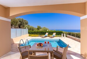 A pool area with a table and chairs, a view of the sea, and a clear sky. - Carme Villas (Rethymno)