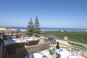 A terrace with wicker furniture, dining tables set for two, and a view of the sea. - Carme Villas (Rethymno)