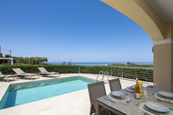 A poolside area with lounge chairs, a dining table set for two, and a view of the sea. - Carme Villas (Rethymno)