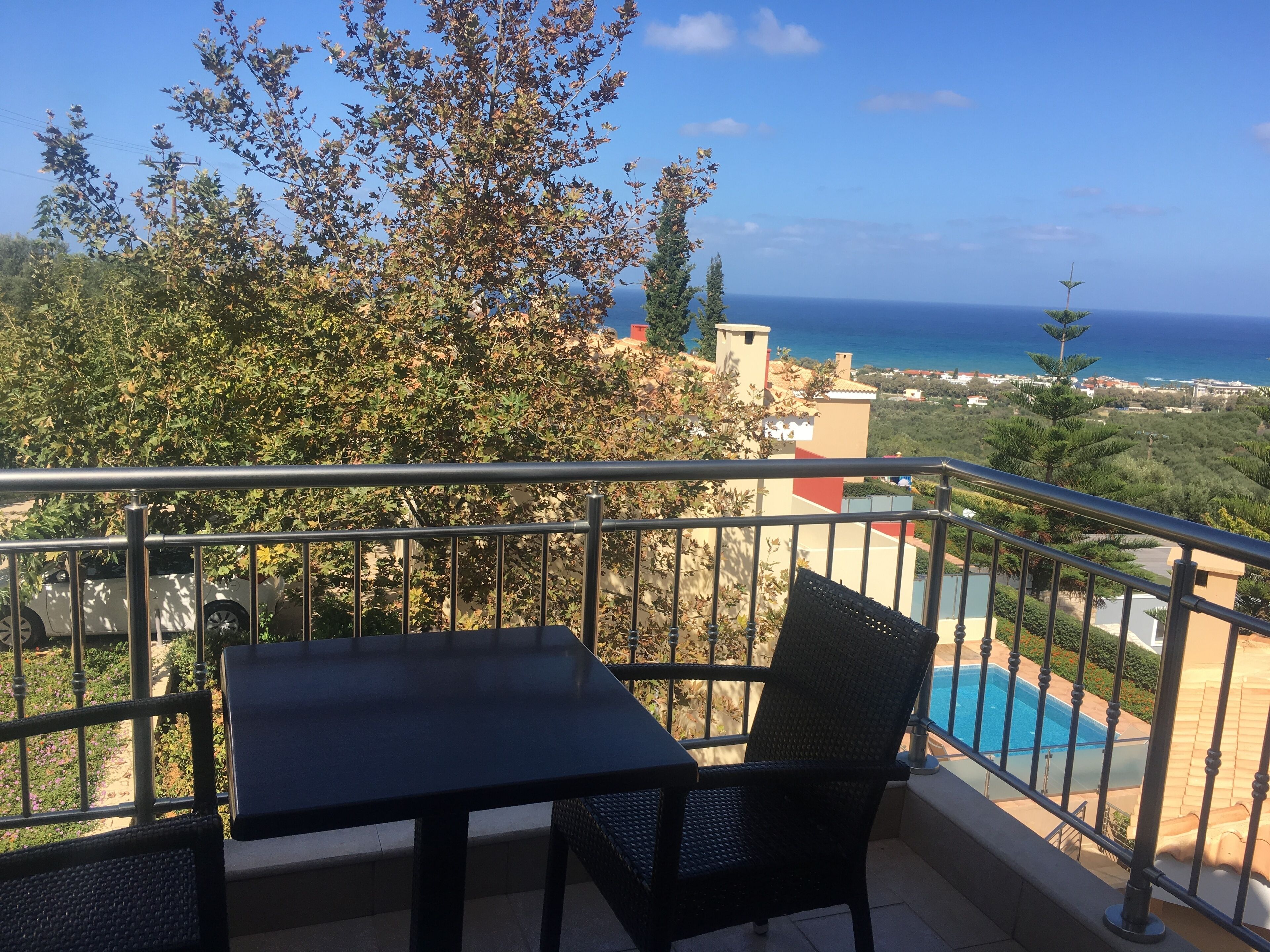 A balcony with a table and chair, overlooking a pool and a view of the sea.