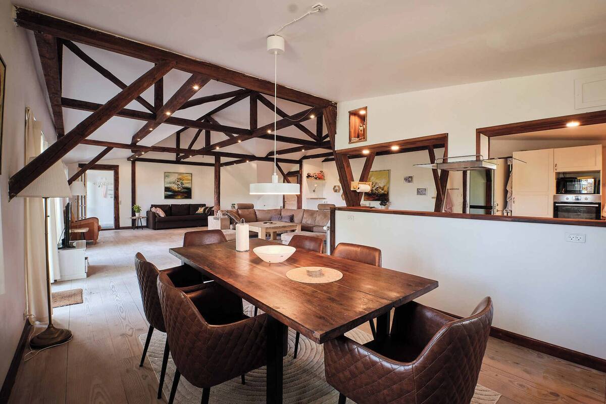 Uma sala de jantar espaçosa com mesa de madeira e cadeiras de couro, teto alto com vigas aparentes e área de cozinha com eletrodomésticos modernos.