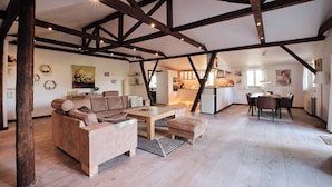 A spacious living area with a high ceiling, exposed wooden beams, a sofa set, a dining table with chairs, and a kitchen in the background. - Askegaarden Apartments (Sandved)