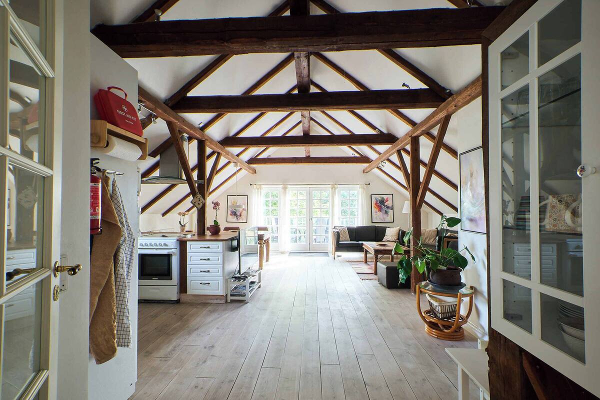 Um quarto amplo no sótão com vigas de madeira aparentes, uma cozinha com fogão e uma sala de jantar com mesa e cadeiras.