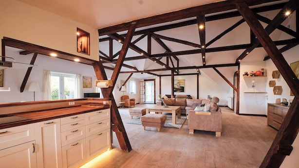 A spacious loft with exposed wooden beams, a kitchen with white cabinetry, and a cozy living area with a sofa and ottoman. - Askegaarden Apartments (Sandved)