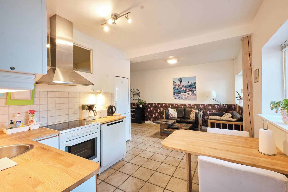 Cozinha compacta com fogão, forno e luminárias modernas. É visível, ao fundo, uma área de refeições com mesa e cadeiras de madeira.