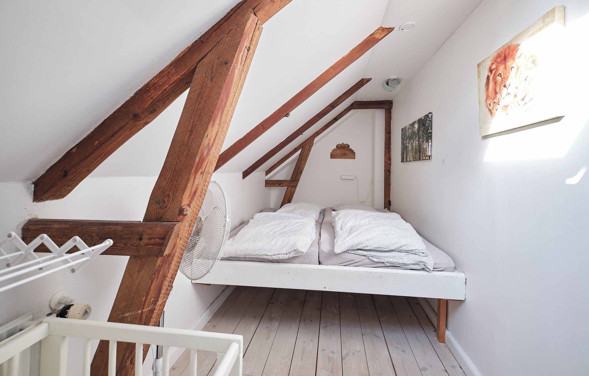 A loft bedroom with a sloped ceiling, wooden beams, a bed with white bedding, and a small wall-mounted shelf.