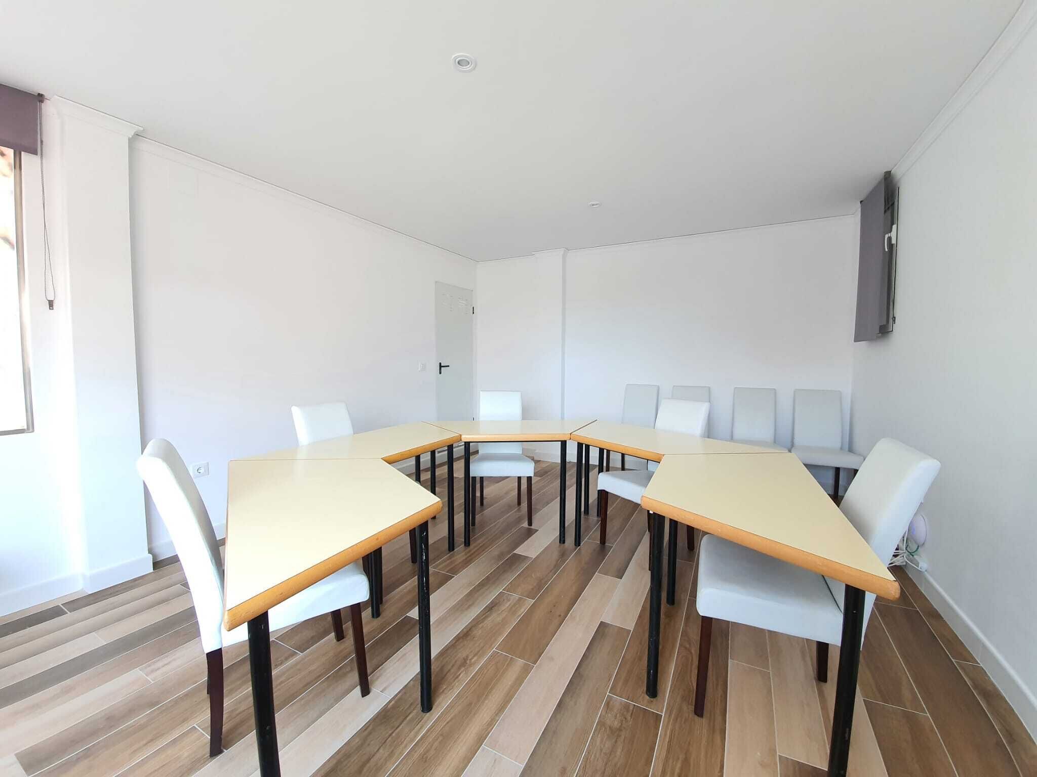 A modern meeting room with a long table and white chairs, wooden flooring, and a white wall with a mounted screen.