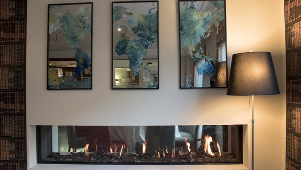 Una chimenea con llamas visibles, tres cuadros abstractos encima y una estantería a la izquierda.