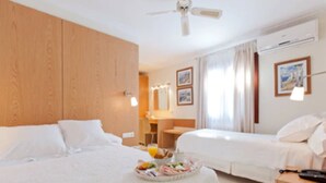 Habitación de hotel con dos camas, cabecero de madera, ventilador de techo, mesita de noche con lámpara y bandeja de frutas.