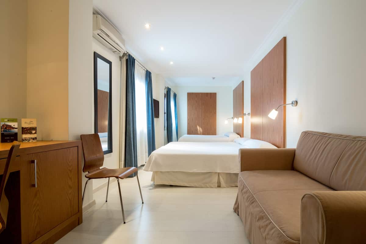 A hotel room with a bed, a desk, a chair, a sofa, and a wall-mounted lamp.