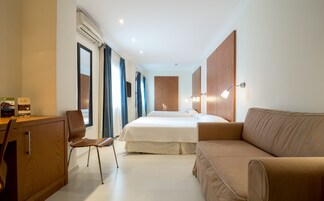 A hotel room with a bed, a desk, a chair, a sofa, and a wall-mounted lamp.