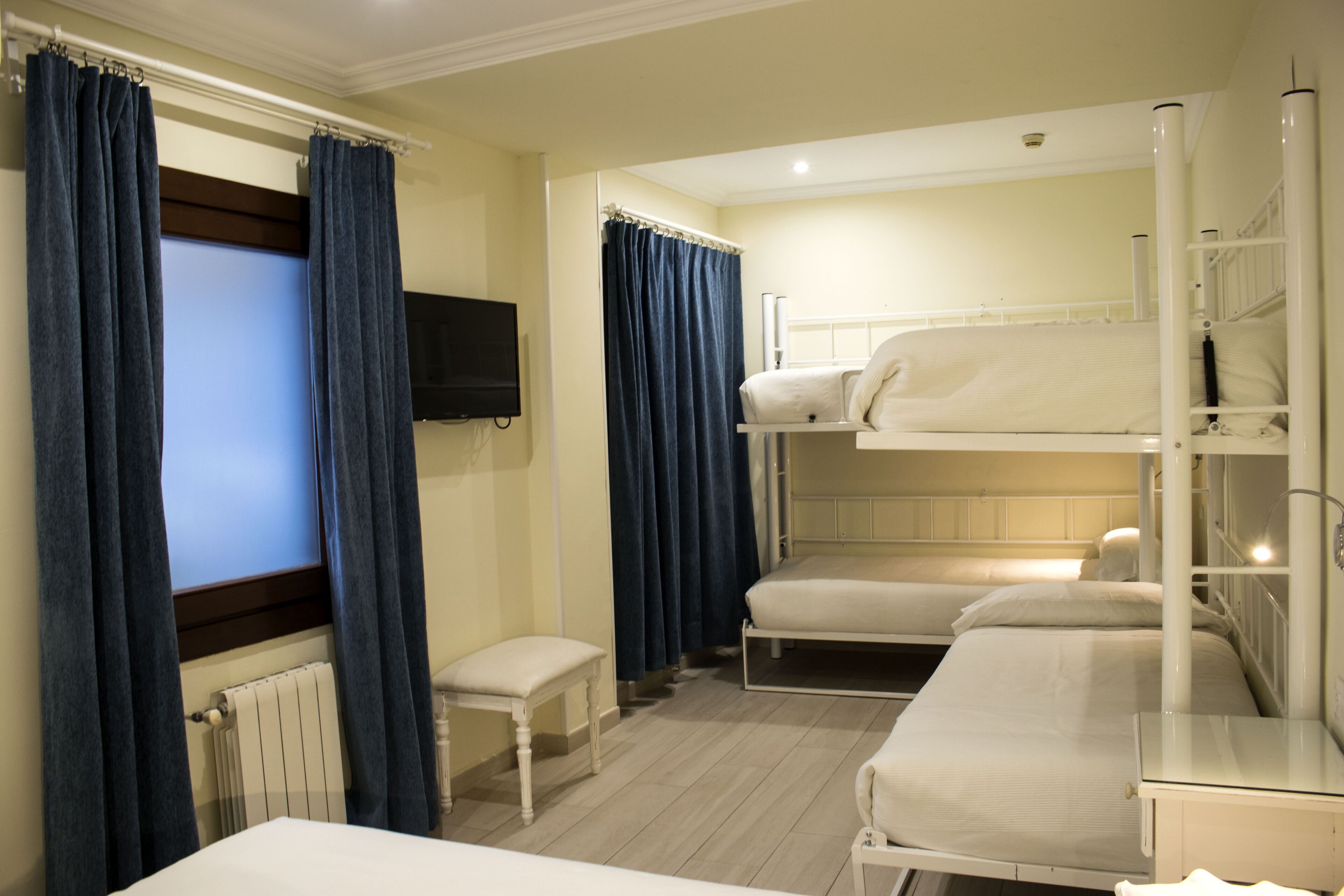 Une chambre d’hôtel avec des lits superposés, une petite table de chevet, une chaise, une fenêtre avec des rideaux et un téléviseur à écran plat fixé au mur.