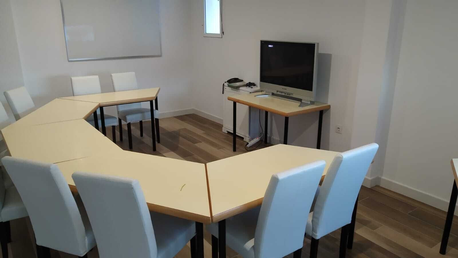 A conference room with a large L-shaped table, six chairs, a television, and a whiteboard on the wall.