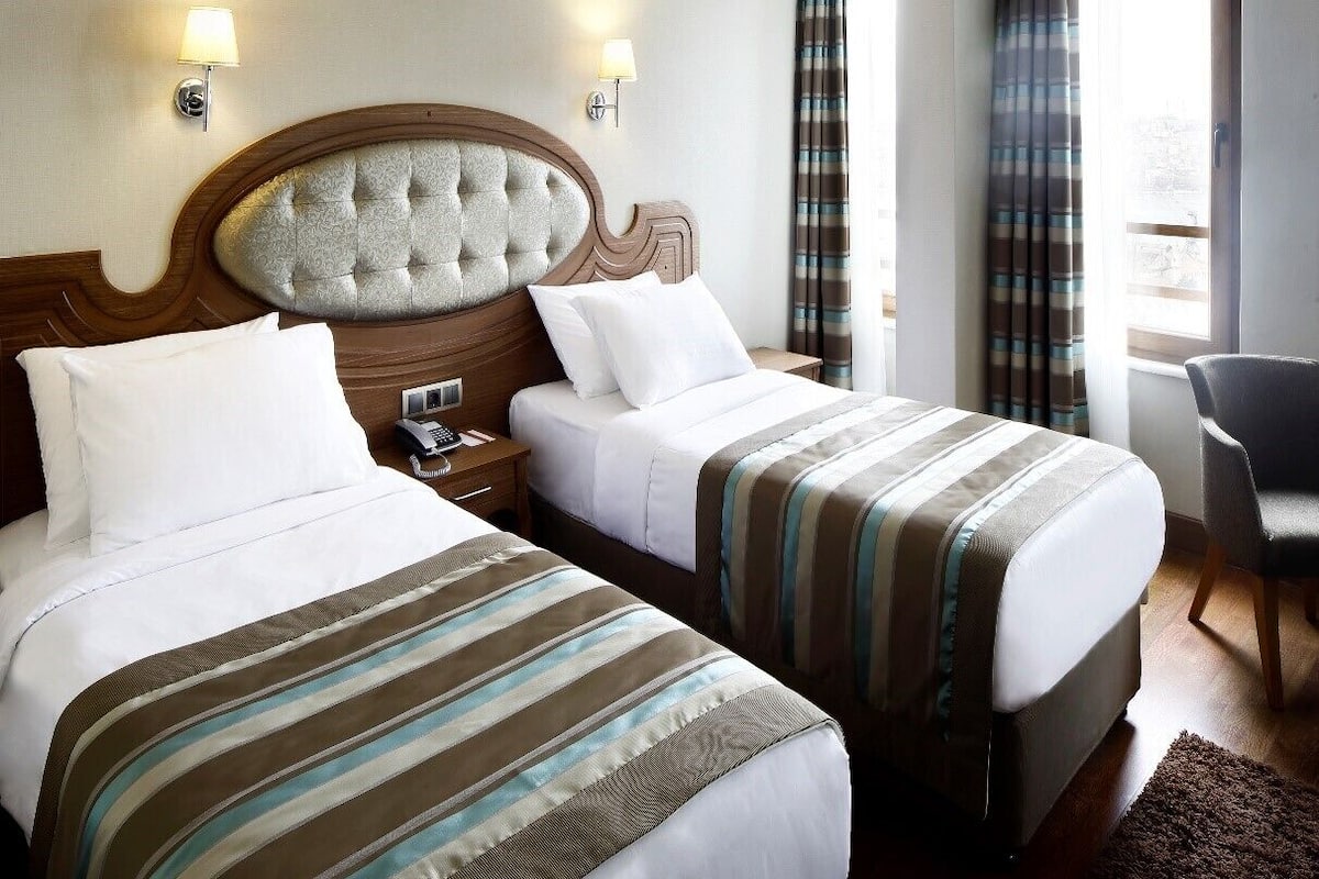 Habitación de hotel con dos camas, cabecero de madera, lámparas de noche, una silla y una ventana con cortinas.