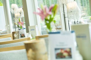 A reception area with a wooden reception desk, a vase with pink flowers, and a large window with a view of greenery. - Gesundheitszentrum Bärenhof Bad Gastein (Bad Gastein)