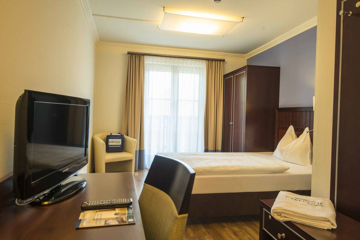 A hotel room with a bed, a desk with a television, a chair, and a bedside table with a remote control and a magazine.