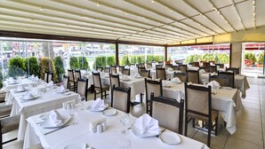 Un restaurant doté de nombreuses tables dressées pour le service, avec un plafond orné de luminaires linéaires et de grandes fenêtres offrant une vue sur la rue et la verdure environnante.
