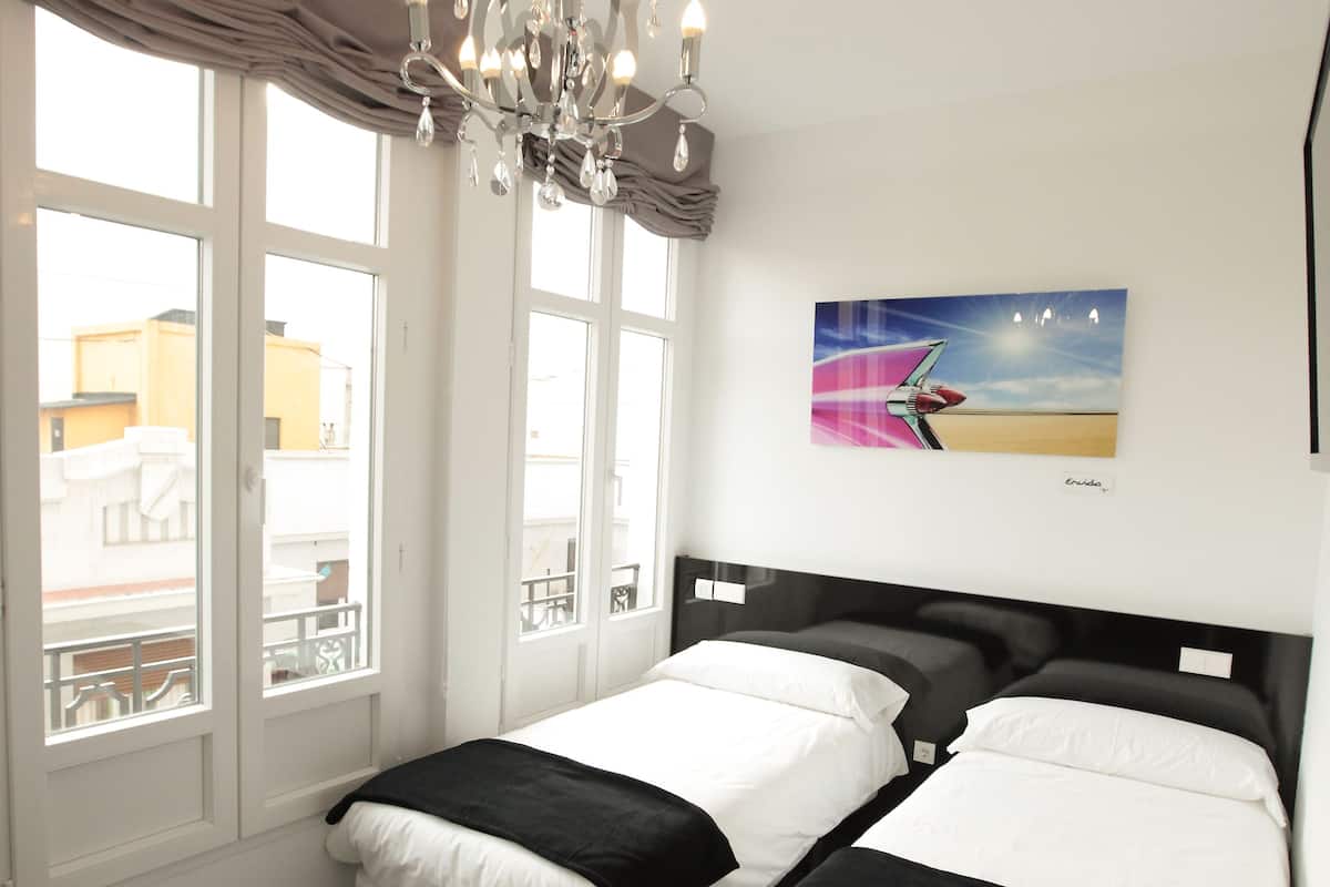 Quarto de hotel com duas camas, um candelabro, janelas amplas e um quadro na parede.