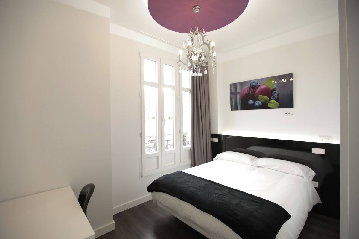 Quarto de hotel moderno com uma cama grande, um candelabro, uma pintura de frutas e vista para o exterior através de uma janela.