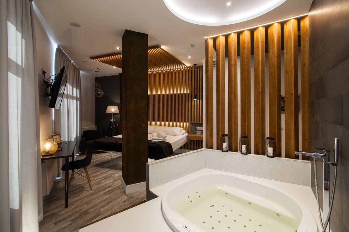 Quarto de hotel moderno com uma banheira grande, uma escrivaninha com cadeira, uma cama e parede revestida de madeira.