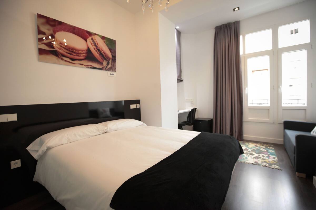 Quarto de hotel com uma cama grande, uma escrivaninha e uma cadeira. Há uma janela com cortinas e um quadro na parede.