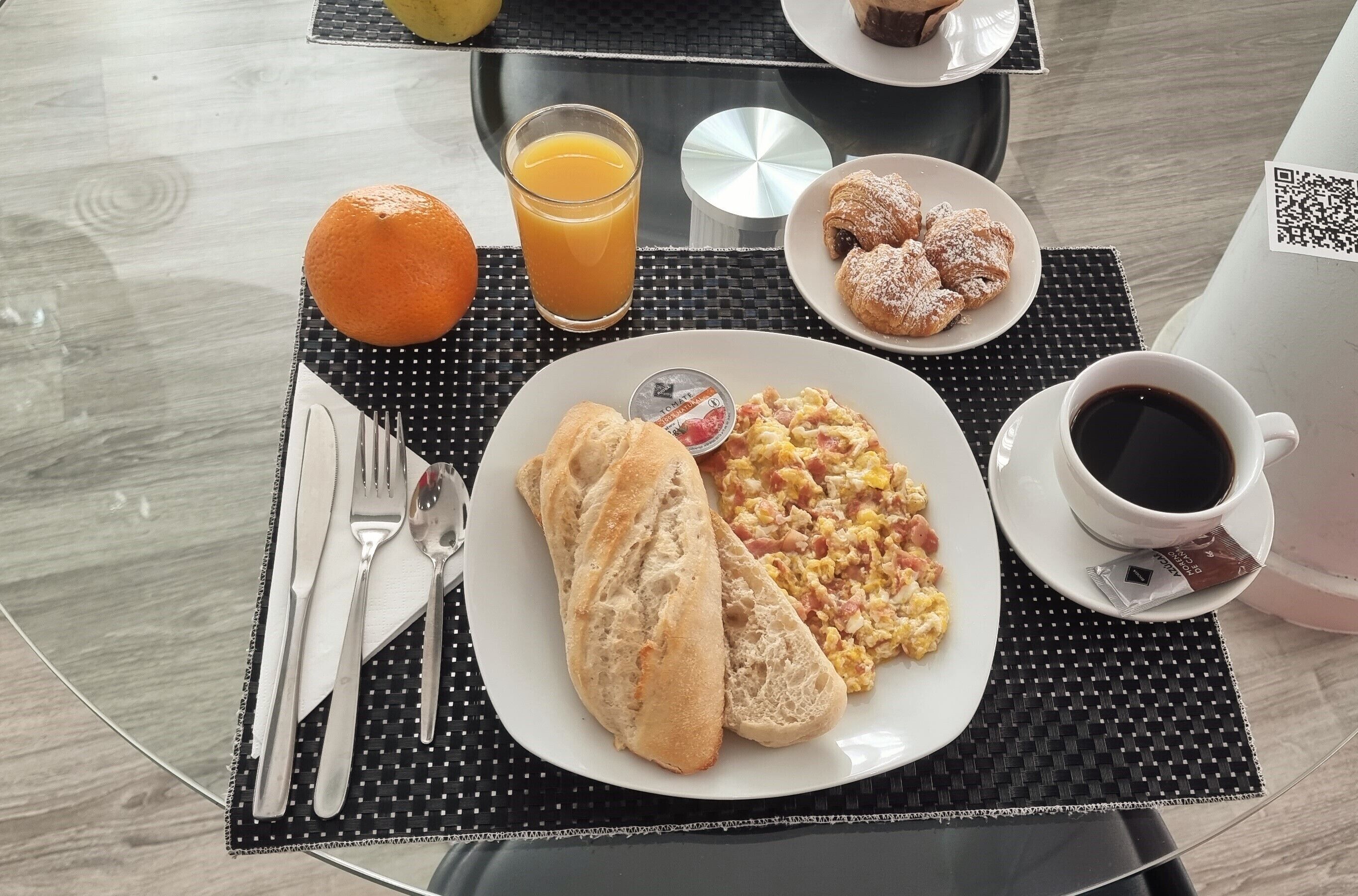 A breakfast setting with croissants, scrambled eggs, orange juice, and coffee on a table with a black placemat.