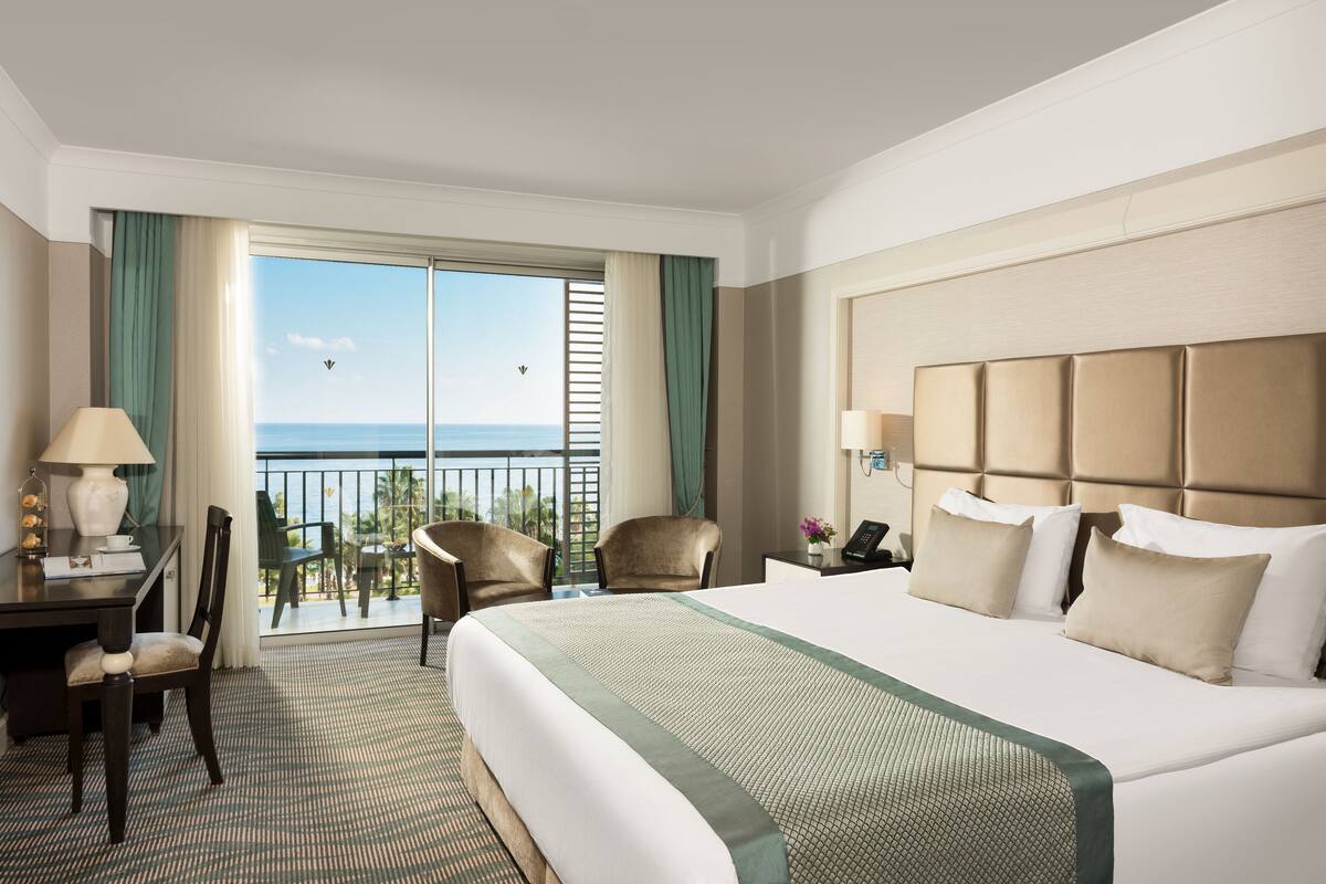 A hotel room with a large bed, a desk with a lamp, two chairs, and a view of the ocean through a sliding door.