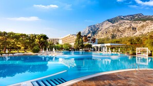 Una gran piscina al aire libre con montañas al fondo, rodeada de exuberante vegetación y edificios modernos.