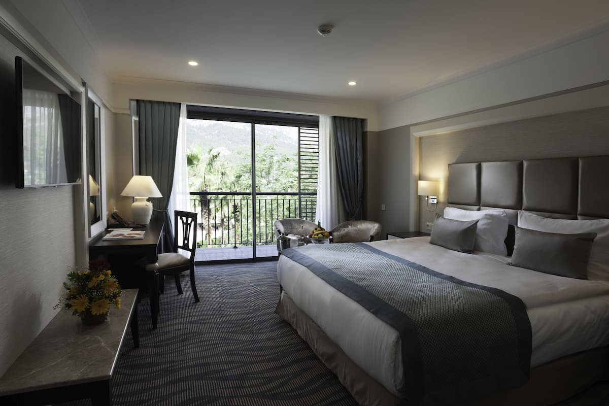 A hotel room with a large bed, a desk with a lamp, a chair, and a balcony with a view of greenery.