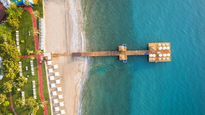 Vista aérea de un muelle frente al mar, con una fila de sombrillas blancas y una zona verde con árboles.
