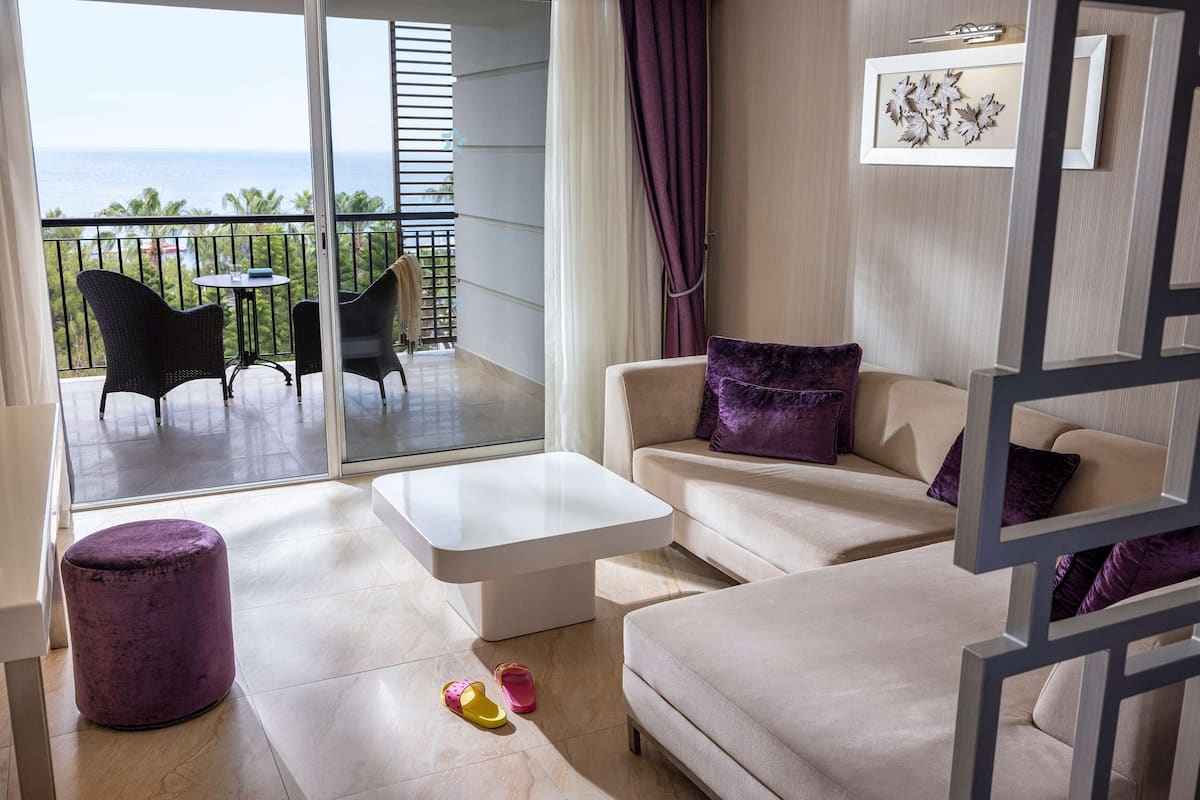 A modern living room with a sofa, ottoman, and coffee table, a balcony with outdoor furniture, and a view of the ocean.