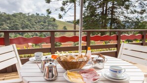 Une table dressée pour le petit-déjeuner, avec du pain, du jambon, du fromage et du café, sur un balcon donnant sur une zone boisée.