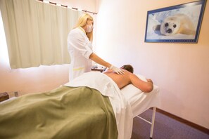 Ein Massageräum mit einer Patientin, die auf einer Liege liegt, einer Therapeutin, die sich um sie kümmert, und einem gerahmten Bild eines Seehunds an der Wand.