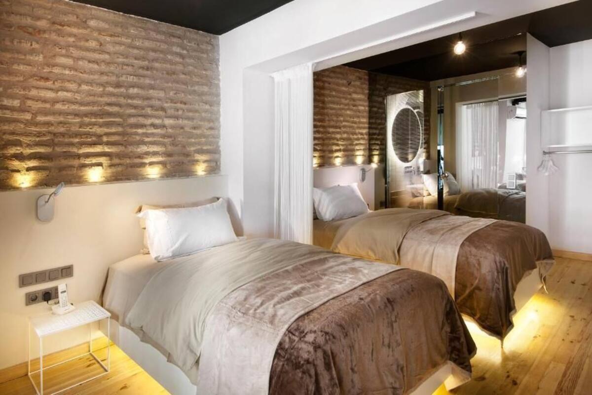 A bedroom with two beds, a brick wall, a mirror, and a bedside table with a lamp.