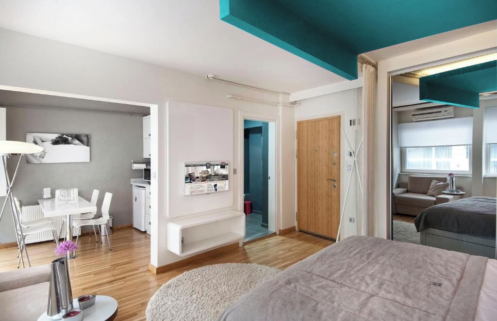 A modern hotel room with a bed, a desk, a chair, a small kitchenette, and a bathroom.