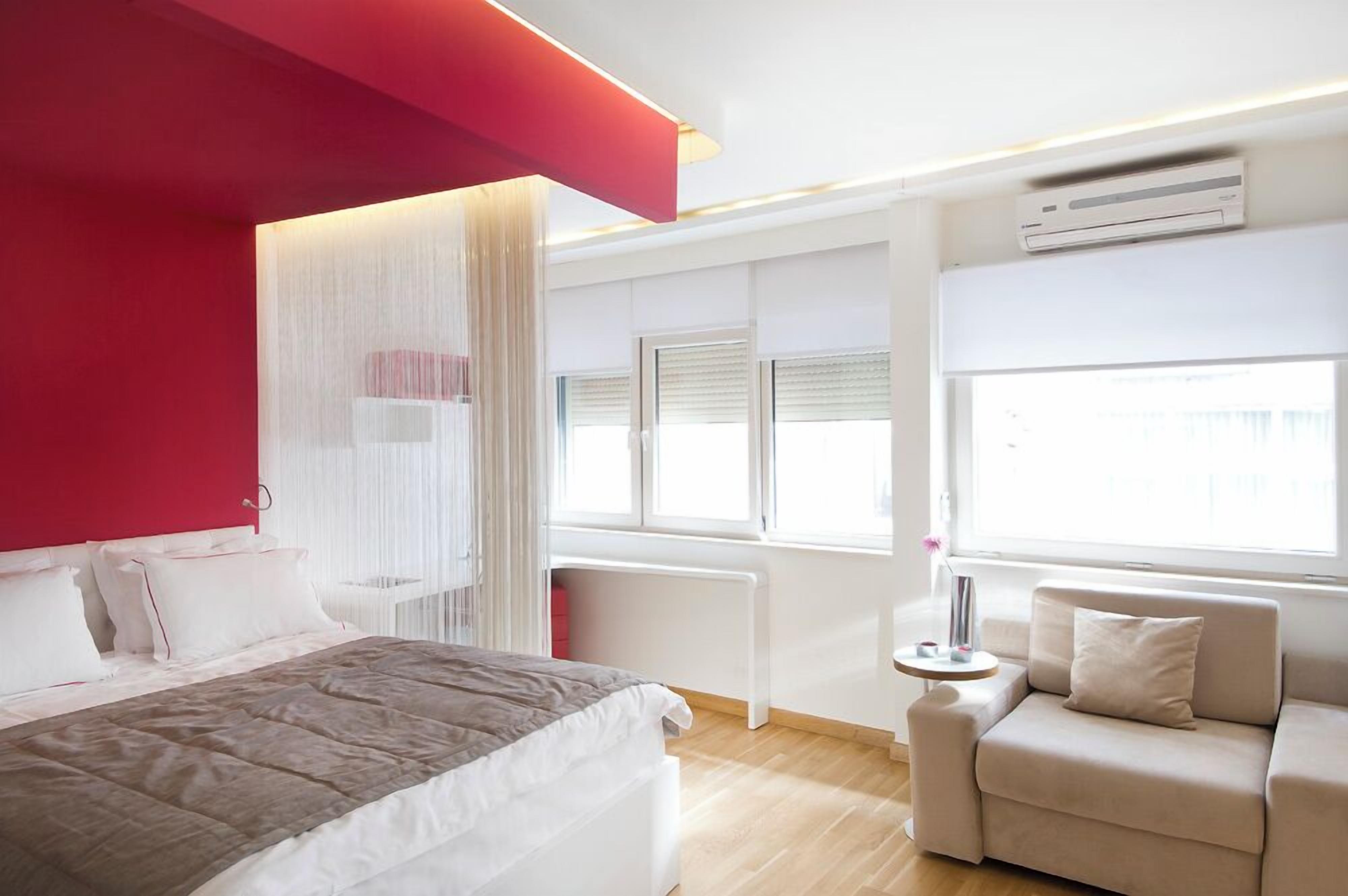 A modern bedroom with a bed, a sofa, a window with blinds, and an air conditioning unit.