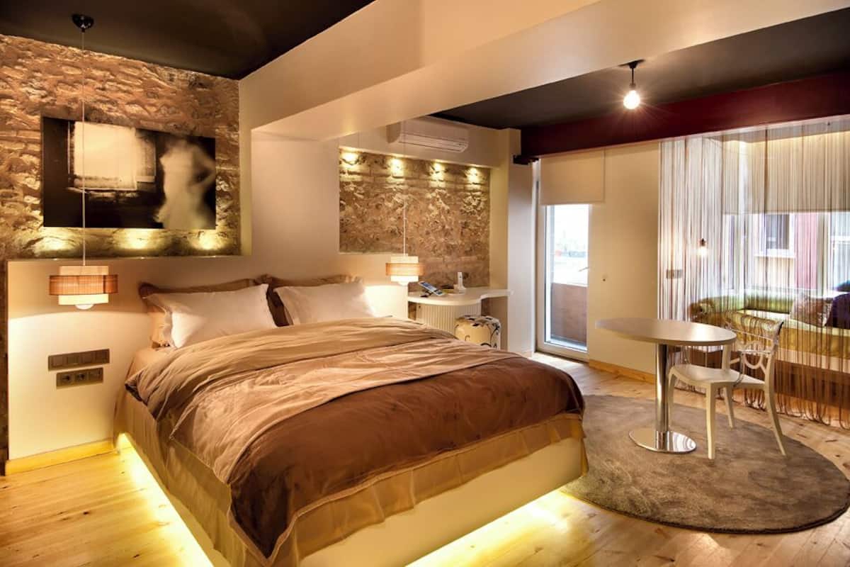A modern bedroom with a stone accent wall, a large bed, bedside tables, a small round table, and a view of the outdoors.