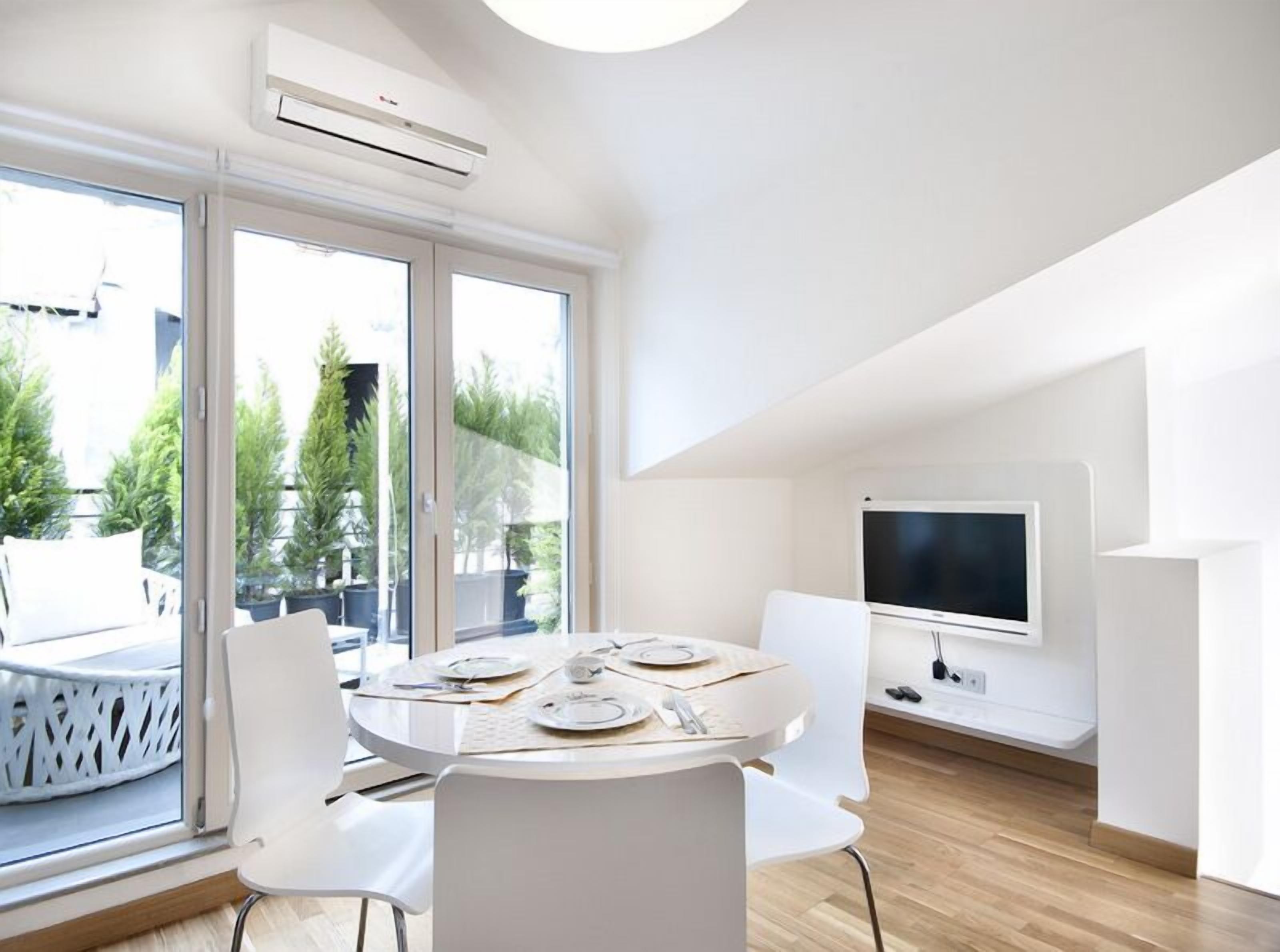 A modern dining area with a round table, white chairs, and a large window with a view of trees and a balcony.