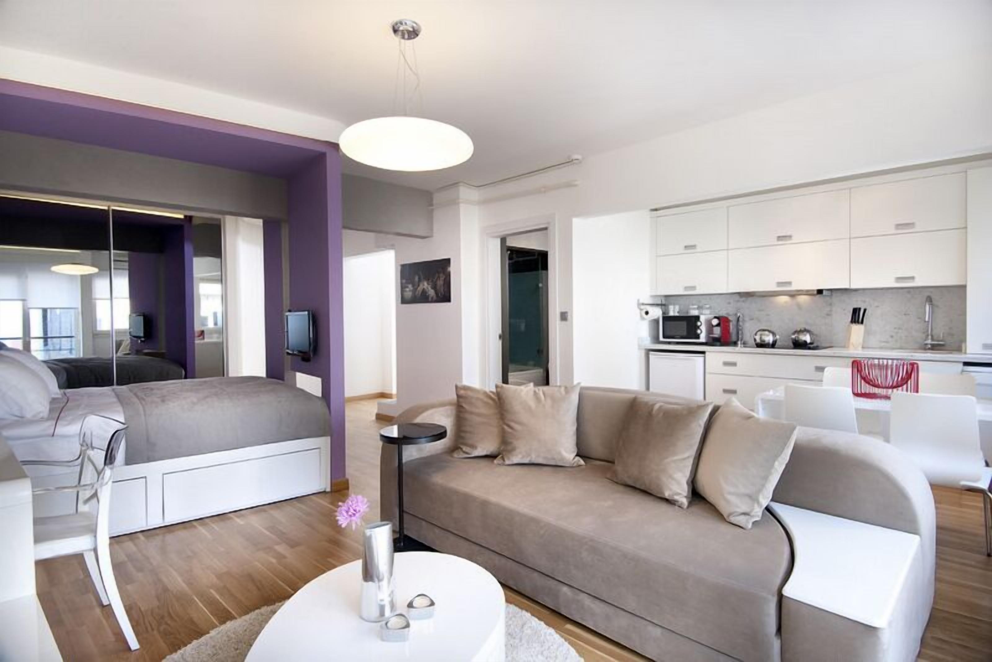 A modern living room with a sofa, a coffee table, and a TV. There is a kitchen area with cabinets and appliances.