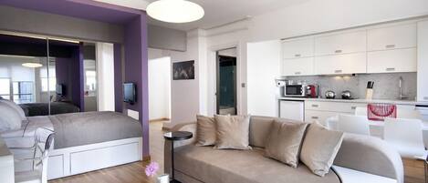 A modern living room with a sofa, a coffee table, and a TV. There is a kitchen area with cabinets and appliances.