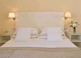 A neatly made double bed with white linens, two bedside lamps, a bedside phone, and a vase with flowers on a small table beside the bed.