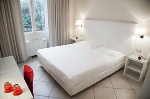 Una camera d'hotel moderna con un grande letto, due comodini con lampade, una sedia rossa e un tavolino con due bicchieri rossi.