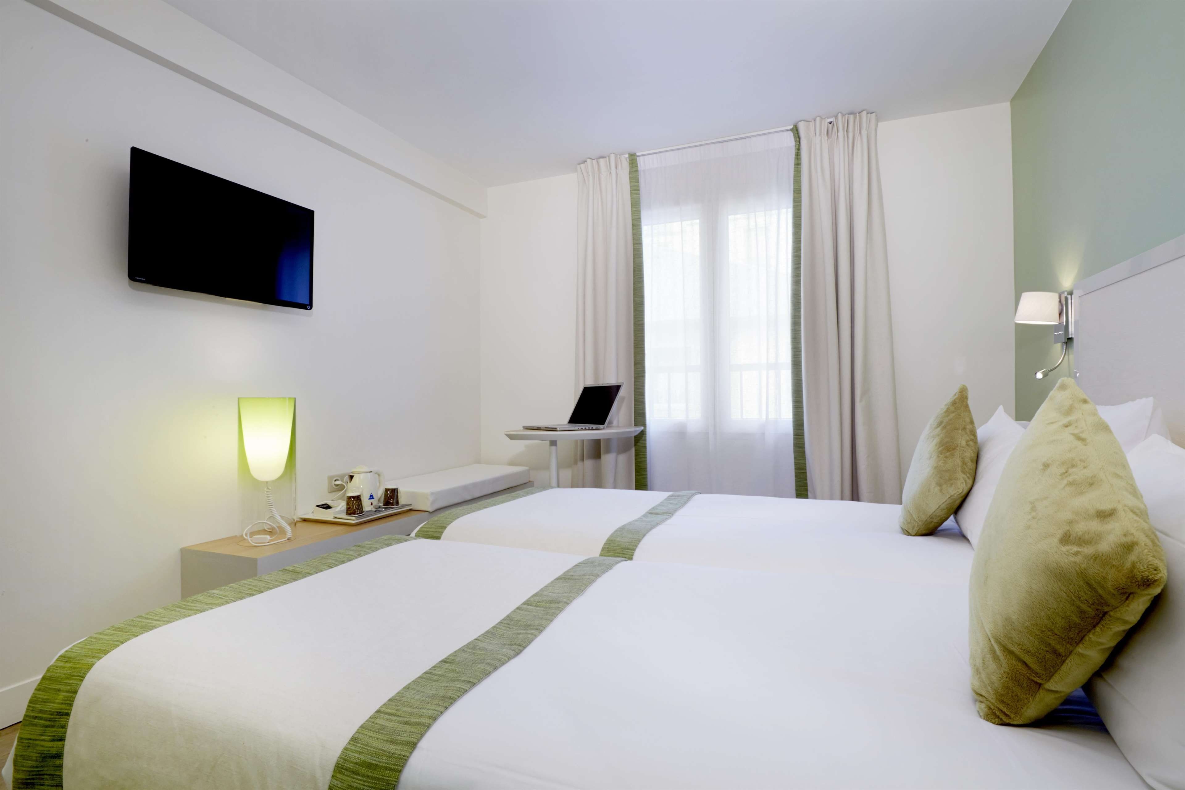 A hotel room with two beds, a flat-screen TV, a desk with a laptop, a bedside table with a lamp, and a window with curtains.