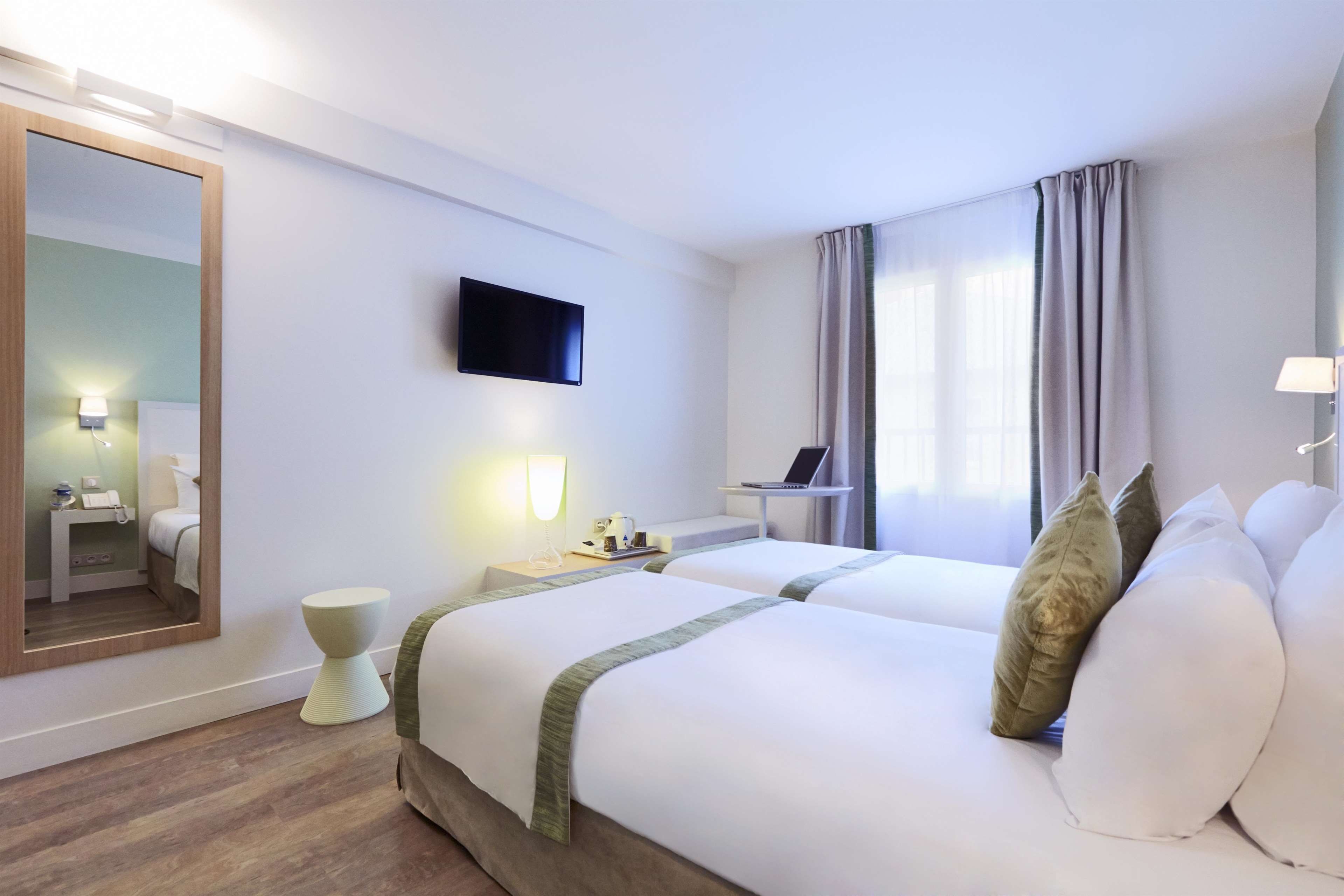 A hotel room with two beds, a flat-screen TV, a bedside table with a lamp, a mirror, and a window with curtains.