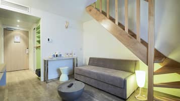 A modern living room with a grey sofa, a round coffee table, a small white side table, a wooden staircase, and a desk with a lamp.
