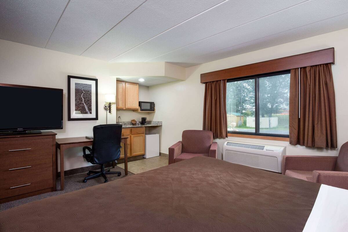 A hotel room with a bed, a desk, a chair, a television, a kitchenette, and a large window with curtains.