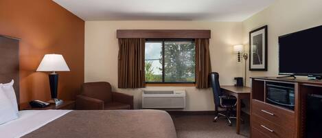 A hotel room with a large bed, a desk with a computer, a chair, a TV, and a window with curtains.