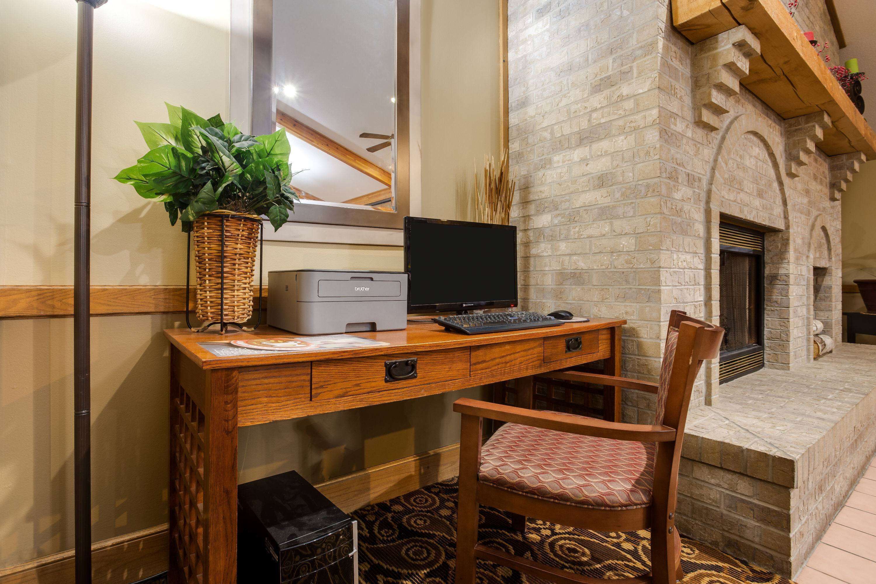 A wooden desk with a computer, a printer, and a plant. There is a chair with a patterned cushion. A fireplace is visible in the background.