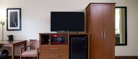 Habitación de hotel compacta con escritorio de madera, televisor, microondas, refrigerador, silla y armario.
