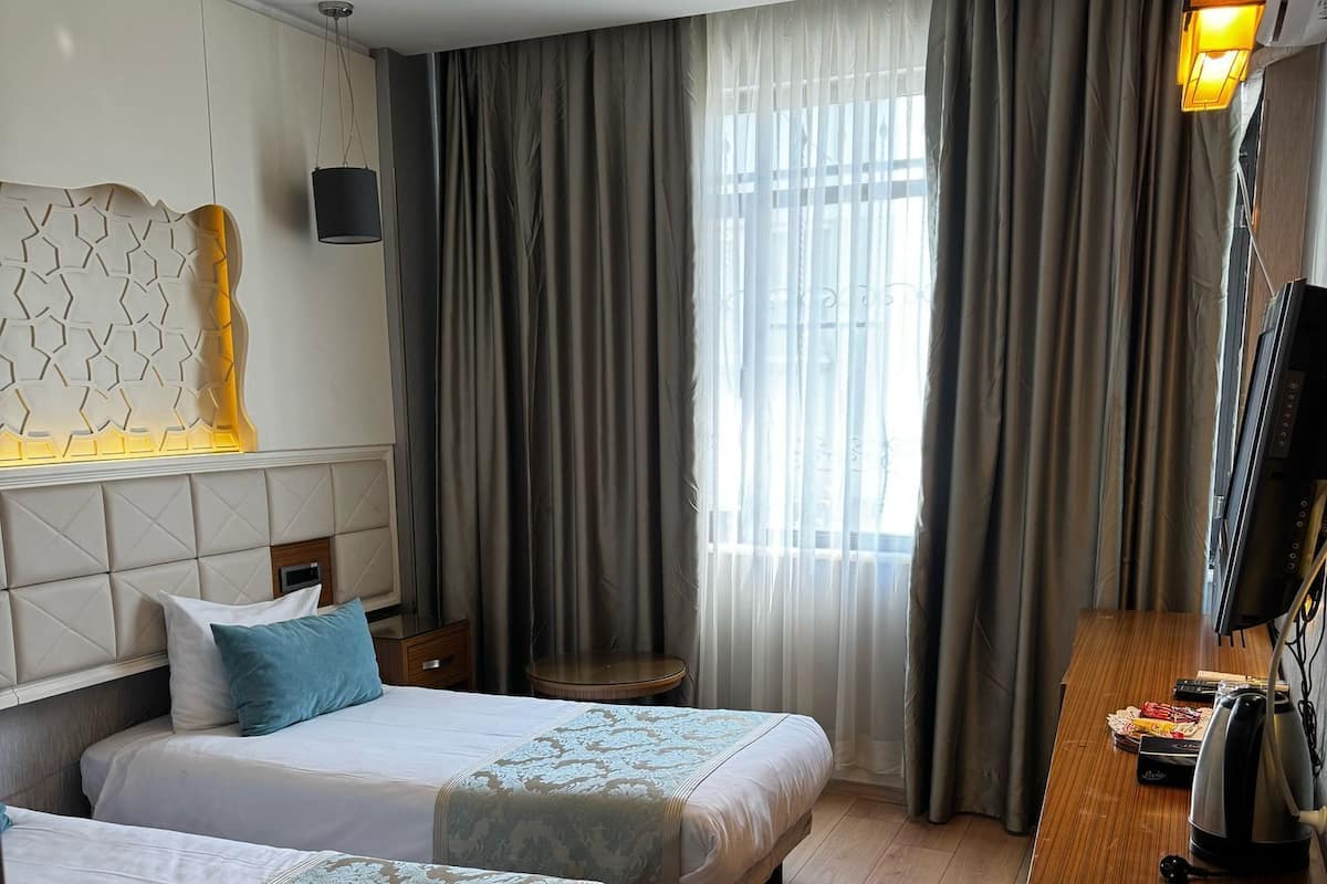 A hotel room with two beds, a wooden desk with a kettle, a flat-screen TV, and a large window with curtains.
